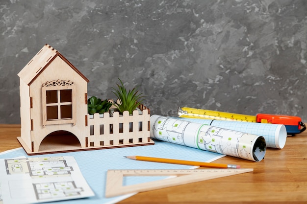 Wooden model house with a white picket fence displayed on a table beside rolled architectural blueprints, a pencil, a triangular ruler, and measuring tools, suggesting home design or construction planning.